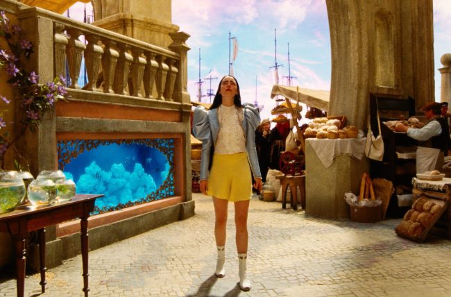 Emma Stone as Bella Baxter wearing yellow shorts, white boots, a ruffle blouse, and a puff sleeve blue jacket.