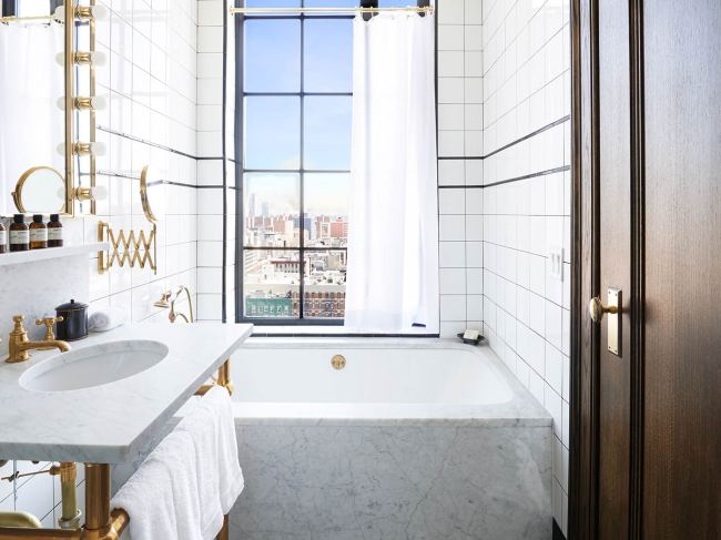 A bathroom at The Ludlow Hotel, featuring a marble bathtub and sink, white-tiled walls, and a window with city views