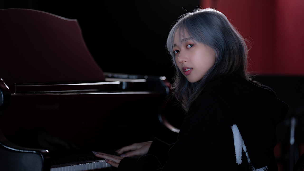 Hannah Bahng, an Asian girl with blue hair wearing a black hoodie playing the piano.