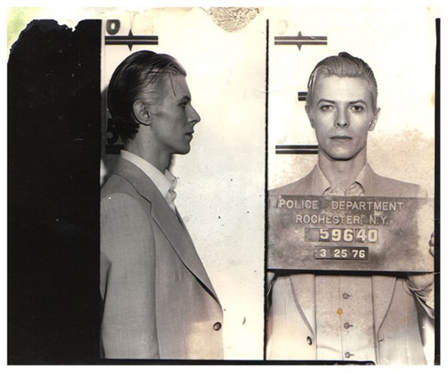 David Bowie poses for his mugshot in a grey suit in Rochester NY.
