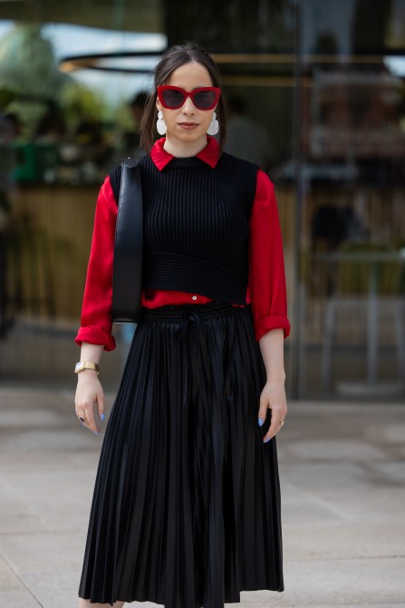 A guest wears red button shirt, black sleeveless knit, pleated skirt, sunglasses, bag outside Marimekko during day four of the Copenhagen Fashion Week (CPHFW) SS25 on August 08, 2024 in Copenhagen, Denmark.