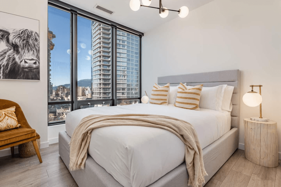 A bright bedroom with a big bed, a ceiling light fixture, a side table, a lamp, and a big window. A downtown Vancouver Airbnb.