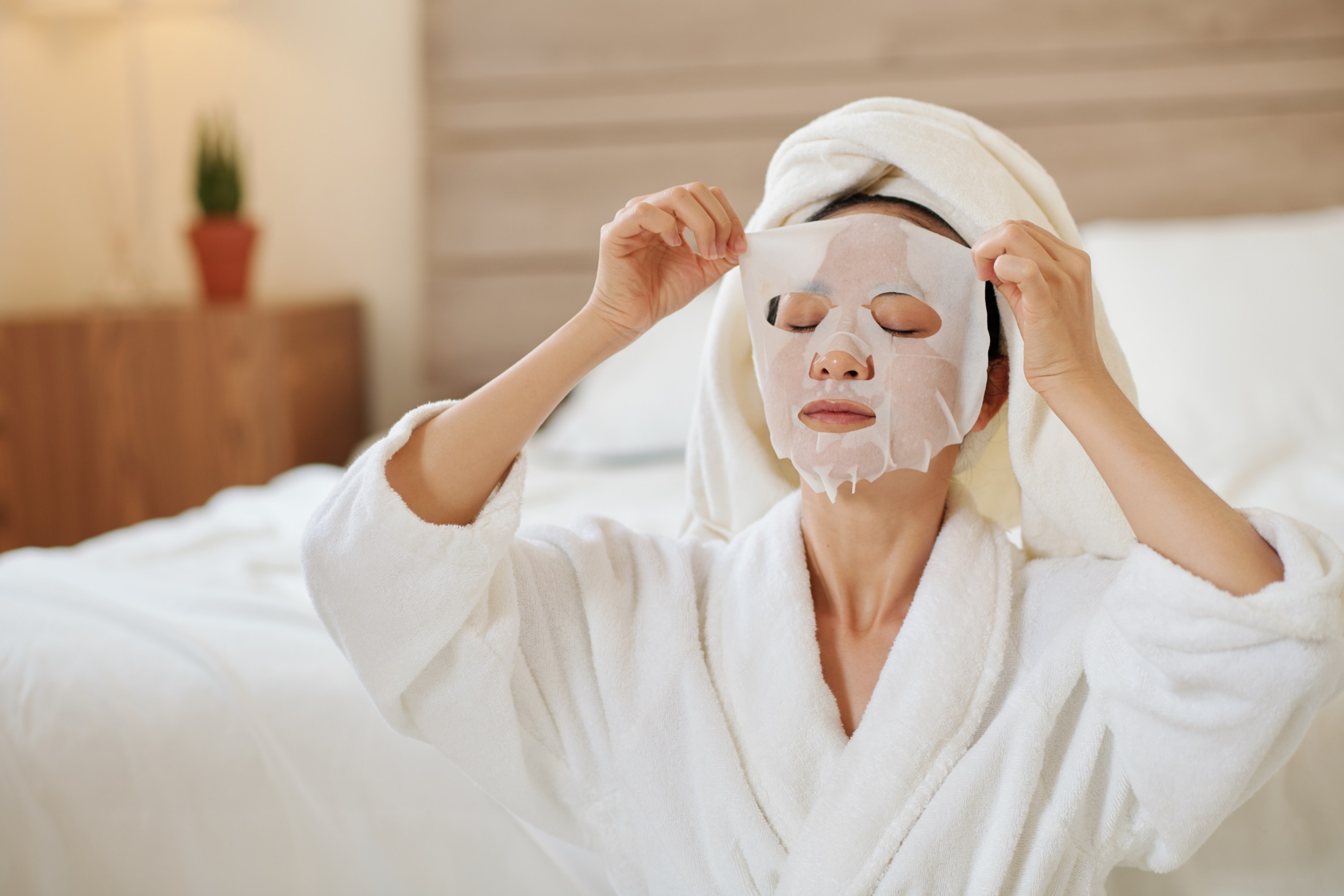 Young woman in bathrobe taking off moisturizing sheet mask