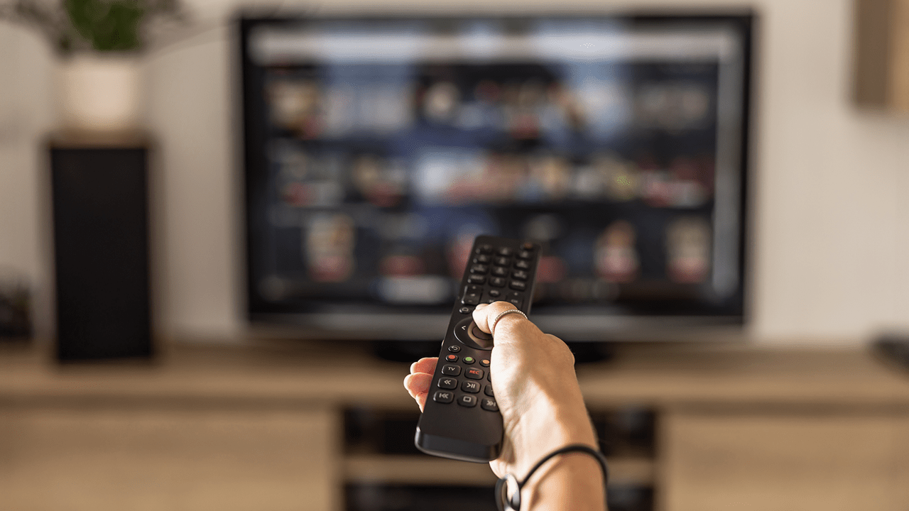 Close up of a person holding a control remote with a television screen on the background. A woman holding a remote control switches programs on the TV in the background.