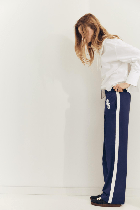 A model wearing the Zara Side Stripe Satin Effect Pants in navy with a white top and black sneakers